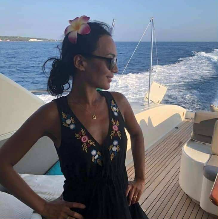 Femme élégante sur un yacht de luxe, portant une robe noire à fleurs et une fleur de frangipanier dans les cheveux, profitant du soleil et de la mer en arrière-plan. Parfait pour un blog de lifestyle, voyage en Méditerranée ou croisière haut de gamme.